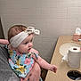 baby, child, headband, floral_dress, arm, hand, adult, holding, bathroom, sink, tiled_wall, countertop, paper_towel_roll, tissue_box, curious, person, indoor, light, skin, tattoo