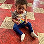 toddler, child, floor, patterned_floor, sitting, crackers, snack, curly_hair, blue_pants, white_socks, colorful_shirt, cartoon_print, indoor, playpen, curious_expression, young_child, person, casual_clothing, looking_up, home