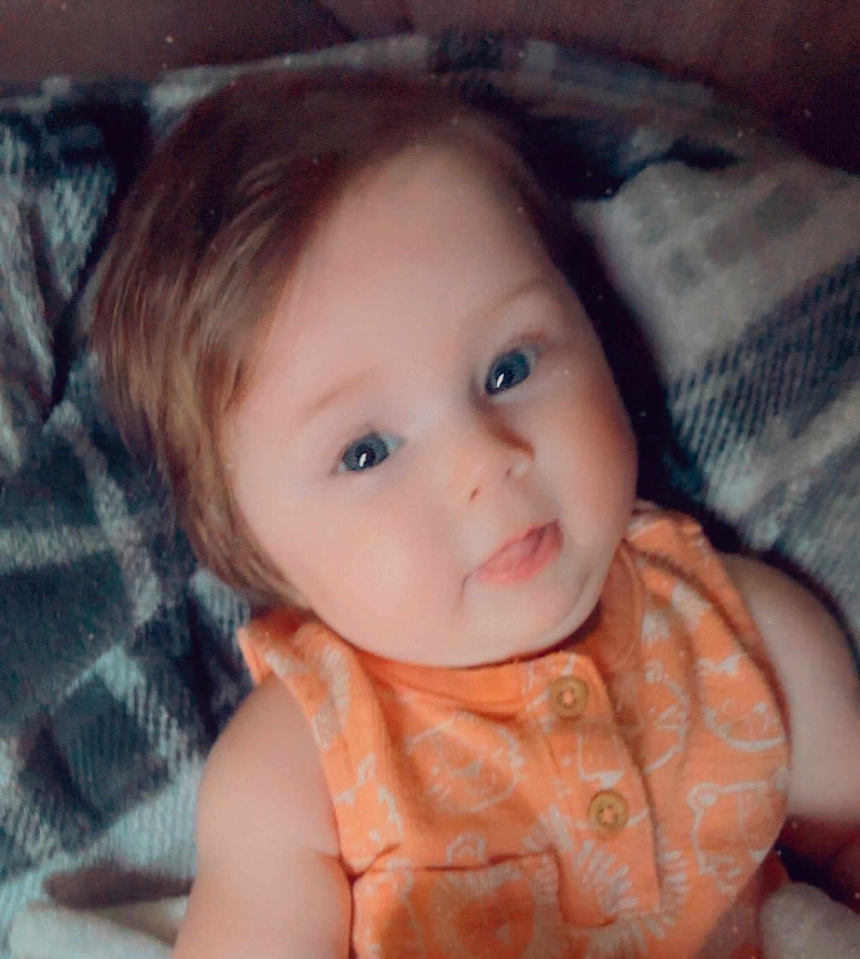 Elianna is registered to the contest to win money with this photo: baby, child, infant, face, blue_eyes, orange_clothing, plaid_blanket, smiling, tongue_out, cute, portrait, closeup, soft_skin, young_child, lying_down, indoors, cozy, happy, person, apparel