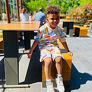 Marcus participe au concours pour gagner de l'argent avec cette photo : child, smiling, curly_hair, colorful_clothing, shorts, sneakers, outdoor, bench, wooden, sunlight, shadow, pavement, park, greenery, people, blurred_background, happy, sitting, daytime, casual
