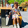 child, smiling, curly_hair, colorful_clothing, shorts, sneakers, outdoor, bench, wooden, sunlight, shadow, pavement, park, greenery, people, blurred_background, happy, sitting, daytime, casual