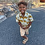 child, smiling, curly_hair, shorts, sandals, tiger_print_shirt, outdoor, paved_road, stone_wall, sunlight, happy, boy, casual_clothing, standing, nature, summer, portrait, daylight, person, cute