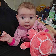 Eliana joined the competition — help win amazing prizes! baby, child, pink_pajamas, plush_toy, turtle_toy, indoor, person, face, hand, seated, furniture, container, sanitizer_bottle, table, toy, clothing, expression, home, infant, cute