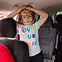 child, face_paint, butterfly, car, tshirt, colorful_text, hands_on_head, seated, adult, window, headrest, seat, inside, curious, person, casual_clothing, blue_eyes, brown_hair, expressionless, daylight