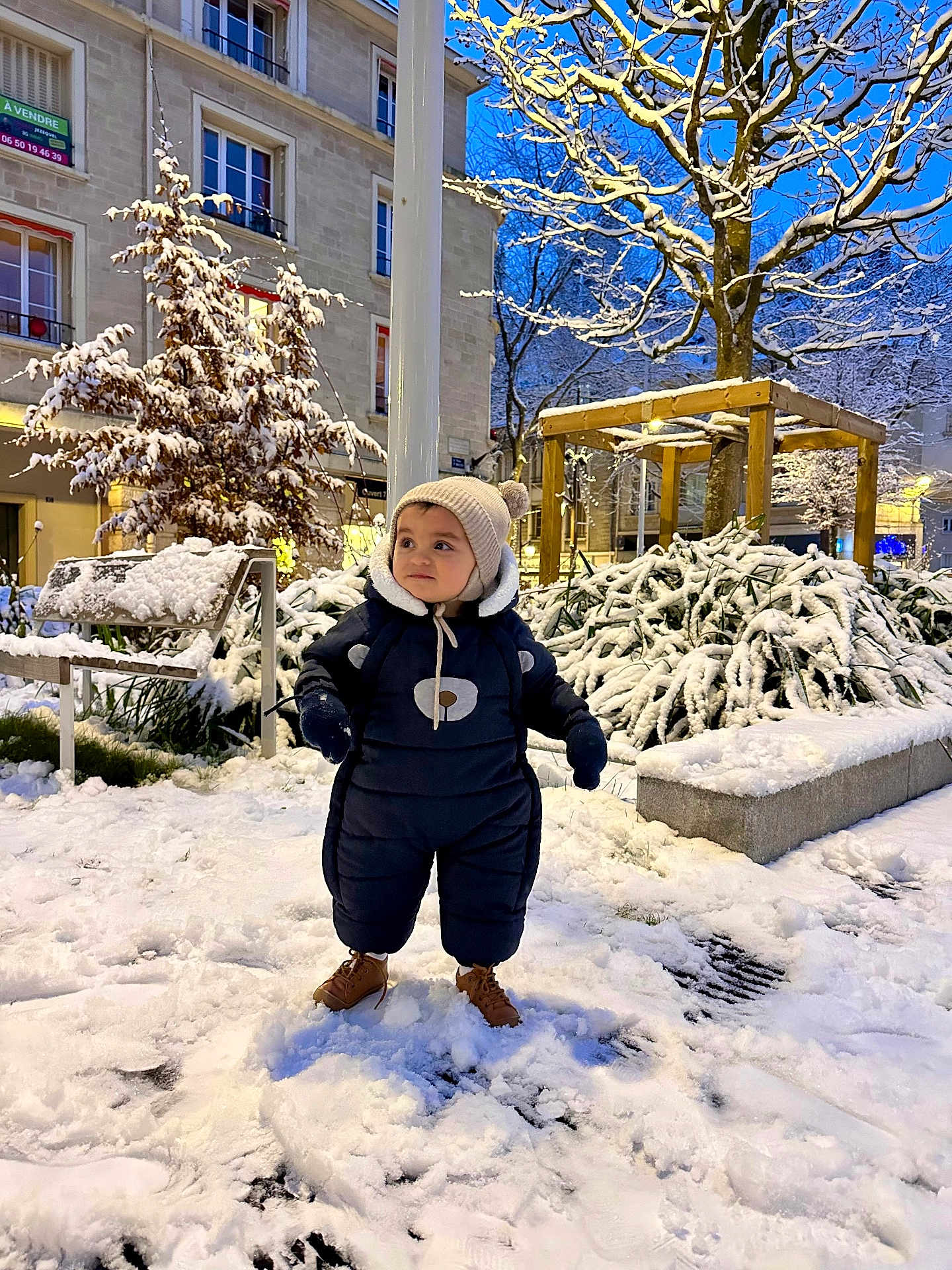 Adam participe au concours pour gagner de l'argent avec cette photo : child, toddler, snow, winter_clothing, hat, boots, snowy_park, trees, bench, urban, evening, street_lights, snow_covered, playful, cute, cold_weather, outdoor, person, warm_clothing, nature