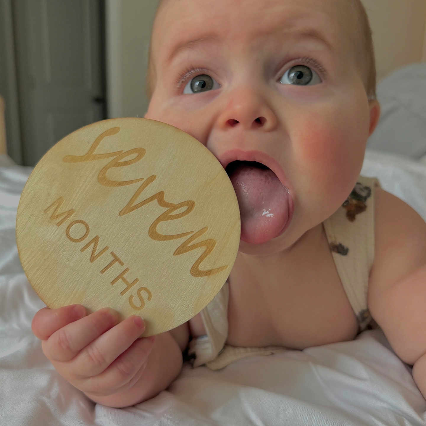 Noah is registered to the contest to win money with this photo: baby, bed, blue_eyes, child, close_up, cute, hand, indoors, infant, light, milestone, playful, portrait, rosy_cheeks, seven_months, skin, tongue, white_bedspread, wooden_plaque, young