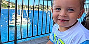 Daniel a rejoint le concours — aidez-le/la à gagner de superbes lots ! adult, background, blue_eyes, child, daylight, hair, happy, outdoor, person, railing, sailboats, seated, shorts, sky, smile, sunny, toddler, tshirt, vacation, water