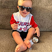 Patrick is registered to the contest to win money with this photo: baby, child, toddler, smiling, sunglasses, baseball_jersey, red_clothing, white_shirt, black_shorts, couch, indoor, seated, barefoot, flashlight, portrait, happy, cute, home, fabric, photography