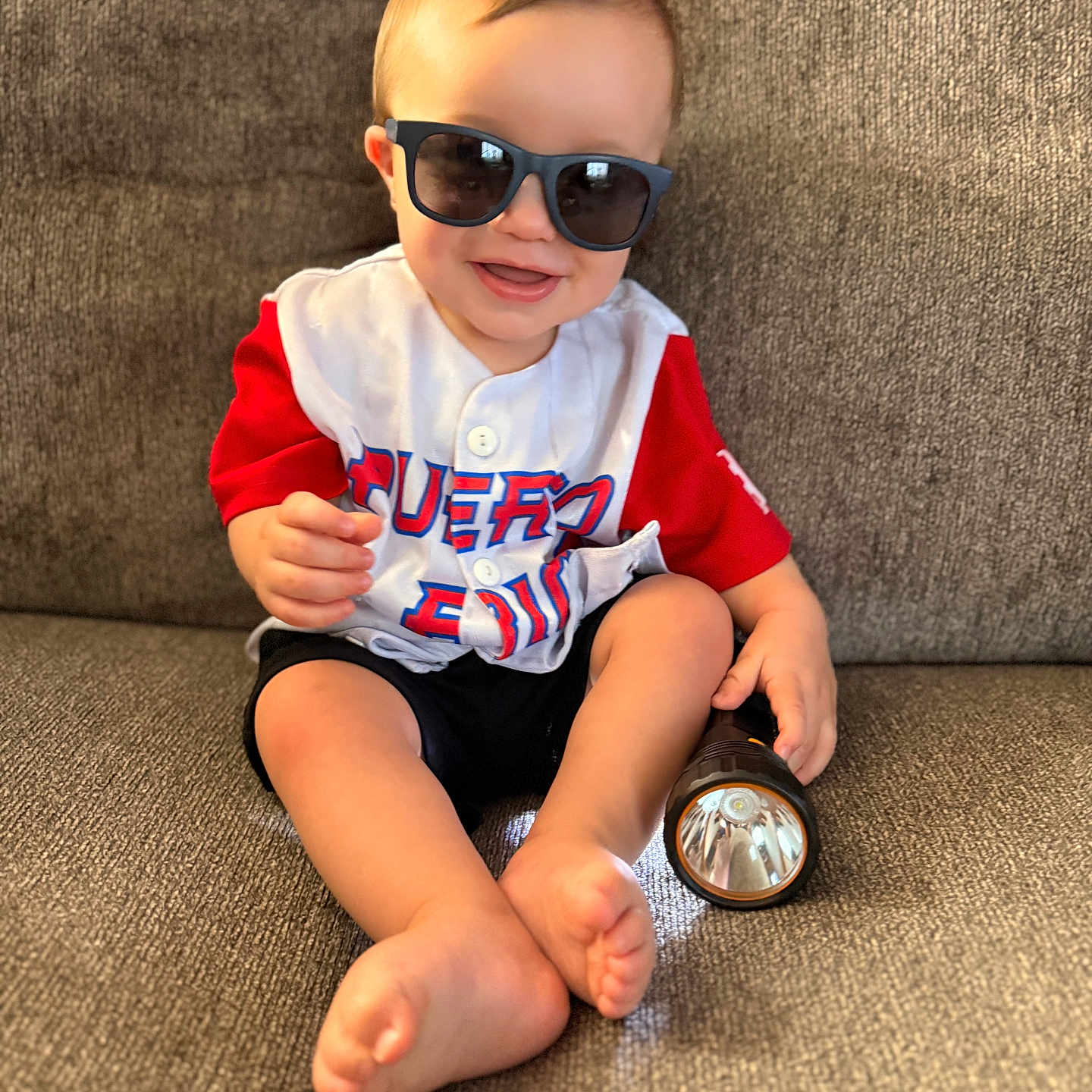 Patrick is registered to the contest to win money with this photo: baby, child, toddler, smiling, sunglasses, baseball_jersey, red_clothing, white_shirt, black_shorts, couch, indoor, seated, barefoot, flashlight, portrait, happy, cute, home, fabric, photography
