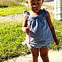 child, toddler, girl, outdoor, grass, sidewalk, blue_dress, white_shoes, hair_buns, daylight, holding_object, street, trees, sunny, casual_clothing, earrings, curly_hair, person, portrait, nature