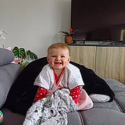 Judith a rejoint le concours — aidez-le/la à gagner de superbes lots ! baby, smiling, polka_dot, blanket, couch, living_room, television, cabinet, plant, red_clothing, happy, child, indoors, furniture, cozy, home, cute, infant, portrait, comfort