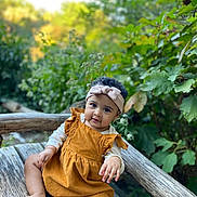Thiare is registered to the contest to win money with this photo: baby, child, headband, dress, wooden_bench, outdoor, nature, greenery, foliage, sitting, cute, portrait, infant, curly_hair, skin, feet, hand, smile, daylight, relaxed
