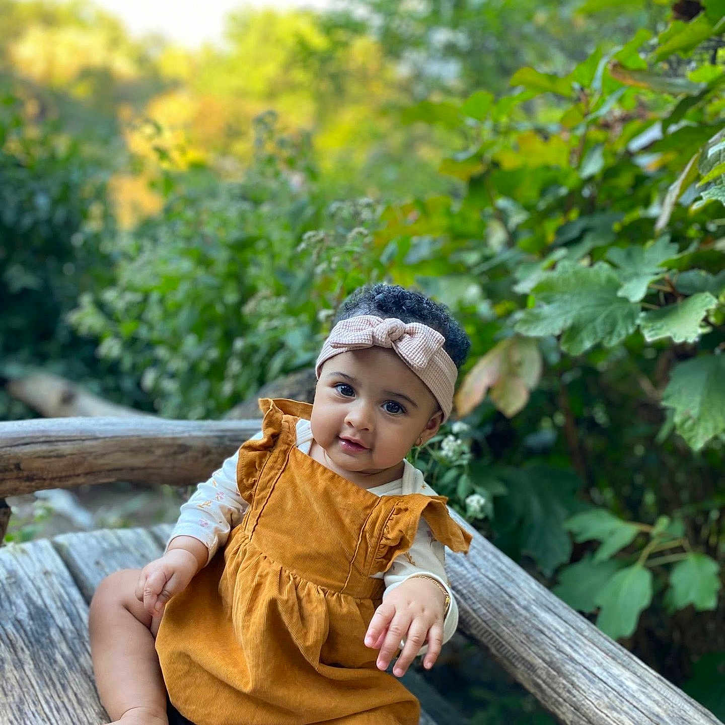 Thiare is registered to the contest to win money with this photo: baby, child, curly_hair, cute, daylight, dress, feet, foliage, greenery, hand, headband, infant, nature, outdoor, portrait, relaxed, sitting, skin, smile, wooden_bench