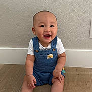 Io Nguyen is registered to the contest to win money with this photo: baby, child, smiling, sitting, blue_overalls, white_shoes, wooden_floor, indoor, happy, cute, person, infant, short_hair, casual_clothing, wall, floor, footwear, young_child, one_person, portrait