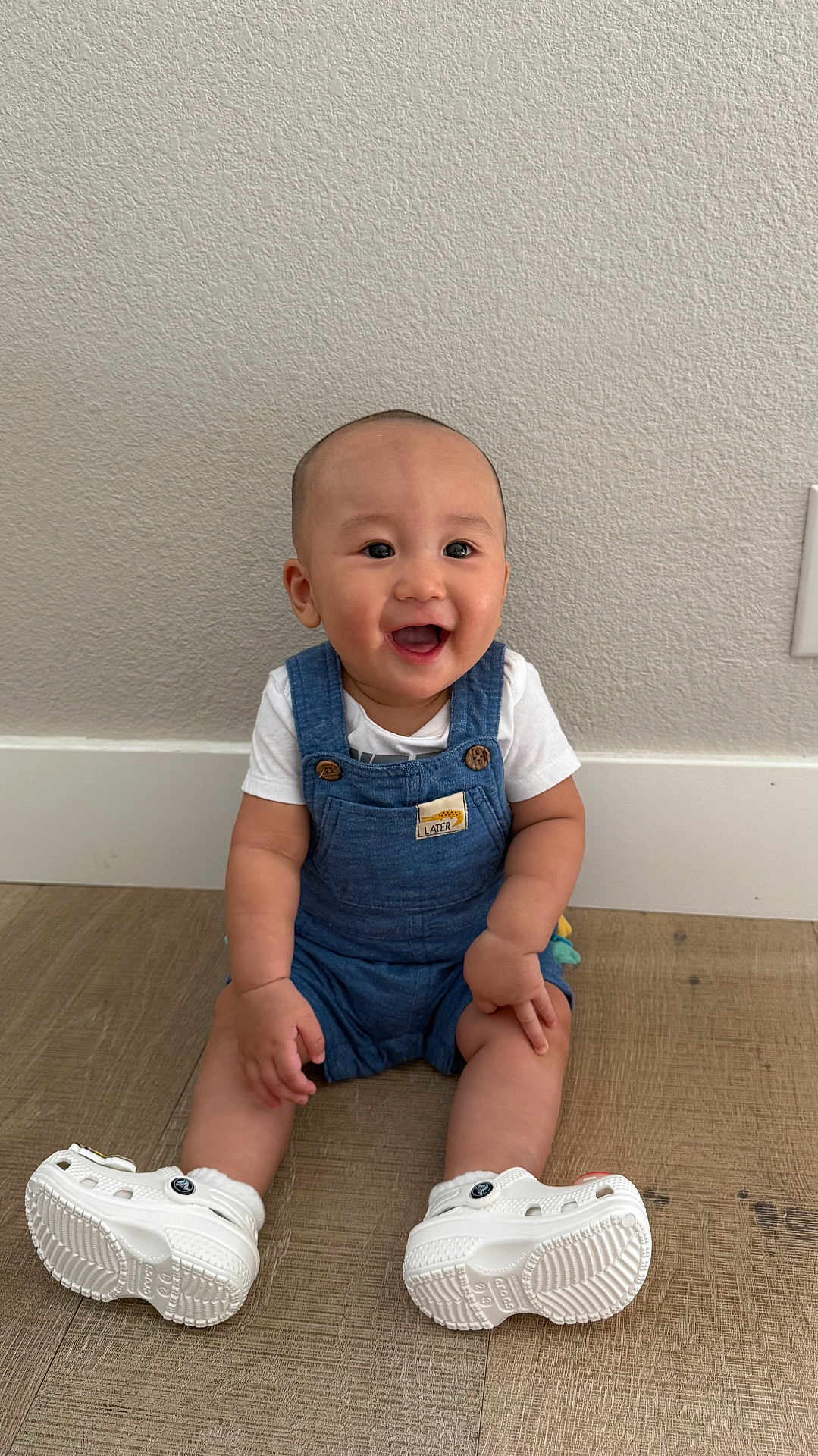 Io Nguyen is registered to the contest to win money with this photo: baby, child, smiling, sitting, blue_overalls, white_shoes, wooden_floor, indoor, happy, cute, person, infant, short_hair, casual_clothing, wall, floor, footwear, young_child, one_person, portrait