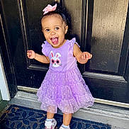 Charleigh is registered to the contest to win money with this photo: child, toddler, girl, smiling, happy, portrait, standing, outdoor, doorway, dress, purple_dress, bunny, hair_bow, pink_bow, sneakers, doormat, home_entrance, curly_hair, joyful, full_body