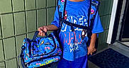 Cameron is registered to the contest to win money with this photo: child, school, backpack, bag, hot_wheels, smiling, portrait, standing, outdoor, school_uniform, blue_clothing, sneakers, toy_cars, doorway, green_wall, sidewalk, school_supplies, happy, school_day, full_body