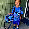 child, school, backpack, bag, hot_wheels, smiling, portrait, standing, outdoor, school_uniform, blue_clothing, sneakers, toy_cars, doorway, green_wall, sidewalk, school_supplies, happy, school_day, full_body
