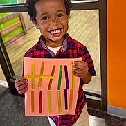 Cameron joined the competition — help win amazing prizes! child, smiling, portrait, indoor, standing, colorful, shirt, plaid_shirt, pants, craft, popsicle_sticks, cardboard, sign, hands, curly_hair, teeth, window, orange_wall, floor, happy