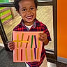 child, smiling, portrait, indoor, standing, colorful, shirt, plaid_shirt, pants, craft, popsicle_sticks, cardboard, sign, hands, curly_hair, teeth, window, orange_wall, floor, happy