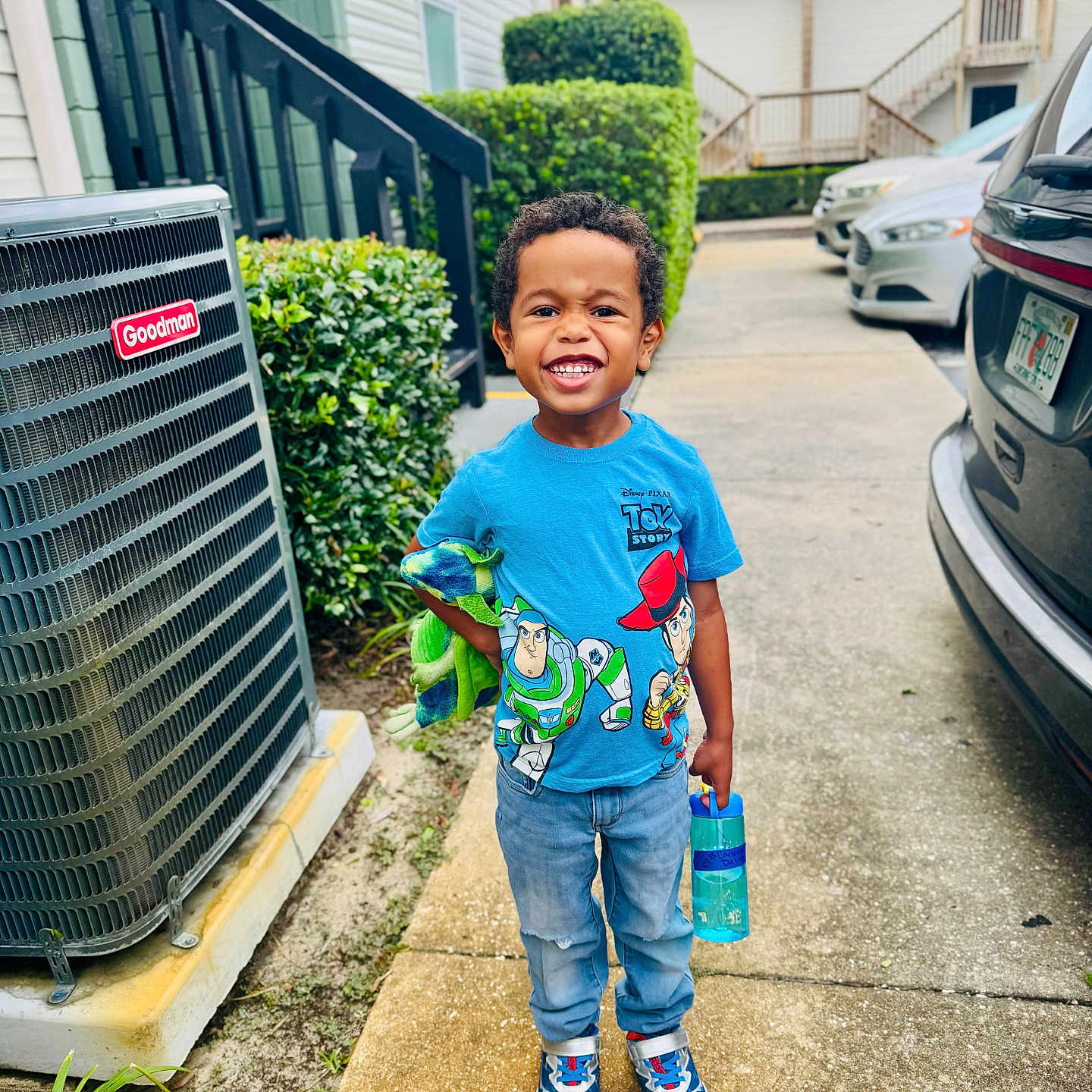 Cameron is registered to the contest to win money with this photo: child, boy, smiling, portrait, outdoor, driveway, residential, air_conditioner, parked_cars, toy, water_bottle, t_shirt, toy_story, blue_clothing, jeans, sneakers, curly_hair, standing, daylight, happy