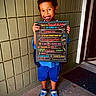 Cameron is registered to the contest to win money with this photo: child, kindergartener, boy, smiling, portrait, school, first_day, back_to_school, sign, chalkboard, doorway, entrance, tile_wall, blue_clothing, backpack, sneakers, standing, outdoor, daylight, happy