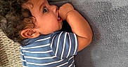 Rhys is registered to the contest to win money with this photo: baby, blue_clothing, candid, carpet, chewing, child, couch, curly_hair, face, hand, home, indoor, infant, photography, portrait, relaxed, sitting, smiling, sofa, striped_outfit
