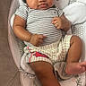 baby, baby_seat, barefoot, brown_skin, child, close_up, cushion, dark_hair, facial_expression, furniture, home, indoor, infant, legs, photography, portrait, shorts, sitting, smiling, striped_shirt
