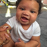 Rhys joined the competition — help win amazing prizes! adult_hand, baby, baby_swing, brown_skin, child, curly_hair, eyes, face, family, floor, happy, home, indoors, infant, onesie, open_mouth, portrait, sitting, smiling, white_clothing