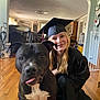 dog, person, graduation_cap, graduation_gown, indoor, wooden_floor, smiling, black_dog, white_chest_marking, tongue_out, living_room, furniture, curtains, cabinet, vacuum_cleaner, pet, happy, portrait, young_adult, home