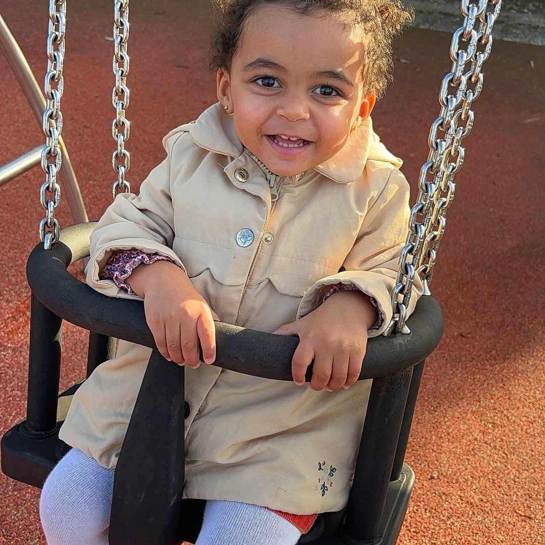 Akia a rejoint le concours — aidez-le/la à gagner de superbes lots ! autumn, bench, casual_clothing, chains, child, curly_hair, footwear, grass, happy, jacket, outdoor, park, person, playground, seat, smiling, sunlight, swing, toddler, toy