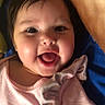 baby, child, smiling, face, pink_shirt, text_on_clothing, dark_hair, cheeks, open_mouth, lying_down, blue_background, brown_background, happy, cute, infant, person, portrait, indoors, clothing, expression