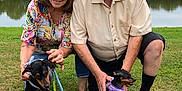 King joined the competition — help win amazing prizes! older_adult, woman, man, dog, leash, grass, pond, trees, outdoor, casual_clothing, cap, glasses, smiling, kneeling, nature, pet, daylight, happy, couple, animal