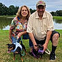 King is registered to the contest to win money with this photo: elderly_couple, woman, man, glasses, cap, dog, dogs, small_dog, leash, grass, pond, outdoor, nature, trees, smile, casual_clothing, shorts, shirt, happy, pets