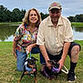 King is registered to the contest to win money with this photo: elderly_couple, outdoor, grass, pond, trees, woman, man, dog, leash, harness, smiling, casual_clothing, cap, glasses, nature, pets, happy, posing, summer, park