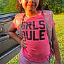 Emma is registered to the contest to win money with this photo: child, girl, pink_shirt, shorts, backpack, smiling, outdoor, grass, car_door, sunlight, curly_hair, confident, standing, hand_on_hip, casual_clothing, nature, daylight, portrait, happy, youth