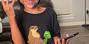 Oliver is registered to the contest to win money with this photo: child, boy, smiling, cap, tshirt, cartoon, phone, indoor, living_room, lamp, couch, wood_floor, window_blinds, happy, casual_clothing, playful, hand_sign, shorts, person, standing