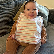 Alzira a rejoint le concours — aidez-le/la à gagner de superbes lots ! baby, smiling, bib, brown_onesie, couch, pillow, soft_fabric, happy, indoor, child, infant, cute, portrait, sitting, face, hand, footwear, clothing, home, young