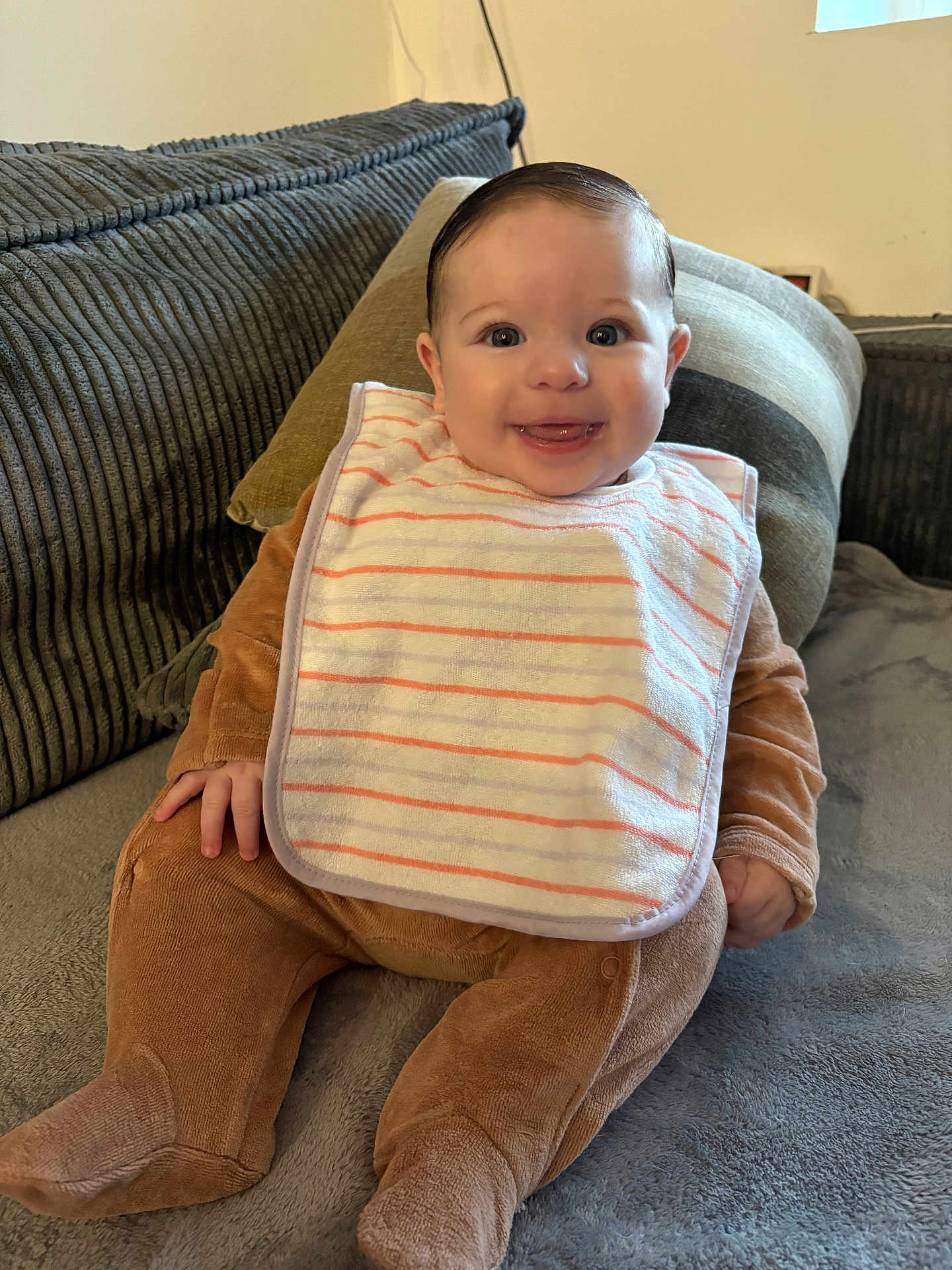 Alzira a rejoint le concours — aidez-le/la à gagner de superbes lots ! baby, smiling, bib, brown_onesie, couch, pillow, soft_fabric, happy, indoor, child, infant, cute, portrait, sitting, face, hand, footwear, clothing, home, young