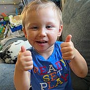 Ronald is registered to the contest to win money with this photo: toddler, child, smiling, thumbs_up, blue_shirt, colorful_shorts, indoor, couch, dog, pet, hair_ponytail, happy, person, furniture, window, blanket, casual_clothing, home, playful, cute