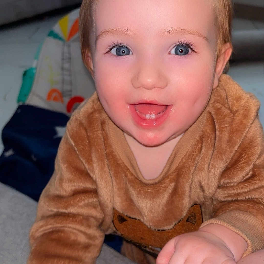 Austin-James is registered to the contest to win money with this photo: baby, blue_eyes, child, closeup, couch, cute, expression, face, fuzzy_clothing, hand, happy, indoor, living_room, person, skin, smiling, sofa, sweater, toddler, toy