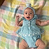 baby, infant, child, portrait, lying_down, blanket, plaid, pastel_colors, blue_outfit, headband, dress, romper, smiling, wide_eyes, raised_arm, outdoorless, close_up, adorable, soft_lighting, textile
