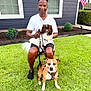 man, dog, pet, grass, yard, house, window, flag, leash, sitting, outdoor, shoes, shorts, white_shirt, fence, shrub, daylight, smiling, chair, portrait