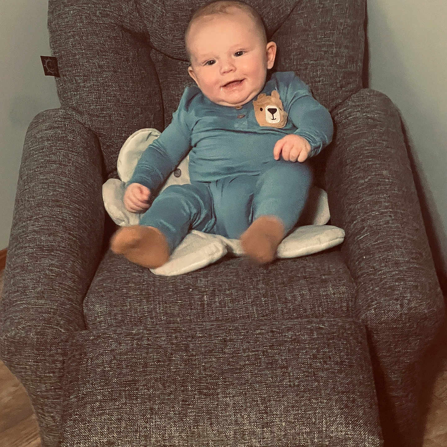 Hunter Jr is registered to the contest to win money with this photo: armchair, baby, bear_patch, blue_onesie, casual, child, cozy, cushion, cute, gray_furniture, happy, indoor, infant, person, plush, portrait, seated, smiling, wooden_floor, young_child