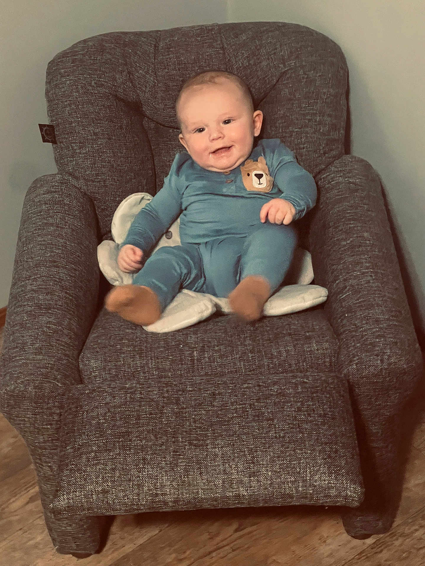 Hunter Jr is registered to the contest to win money with this photo: baby, child, smiling, blue_onesie, bear_patch, armchair, gray_furniture, cushion, indoor, wooden_floor, person, seated, happy, cute, infant, young_child, cozy, plush, portrait, casual