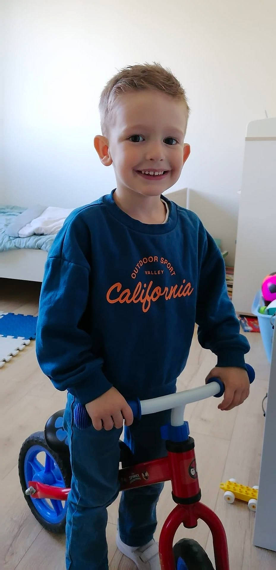 Alexis participe au concours pour gagner de l'argent avec cette photo : balance_bike, bed, blue_sweatshirt, boy, casual_clothing, child, floor, footwear, furniture, hand, happy, indoor, pants, person, play, room, smile, toy, wooden_floor, young