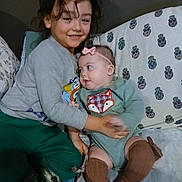 Shi is registered to the contest to win money with this photo: child, baby, sibling, hug, smile, couch, blanket, headband, long_sleeve_shirt, onesie, knee_high_socks, patterned_pillow, indoor, family, cute, toddler, happy, cozy, casual_clothing, portrait