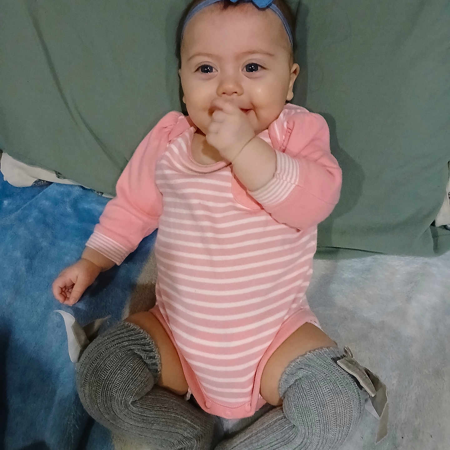 Shi is registered to the contest to win money with this photo: baby, blanket, bow, child, cute, gray_socks, hand, happy, headband, human, indoors, infant, lying_down, onesie, person, pink_clothing, smiling, socks, striped_clothing, toddler