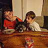 boy, child, dog, table, plate, glass, indoor, furniture, curtain, wall, lamp, casual, family, pet, affection, wood, sitting, smiling, face, clothing