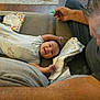 Wesley is registered to the contest to win money with this photo: baby, smiling, couch, person, adult, tissue_box, remote_control, pacifier, blanket, wood_floor, home, indoor, relaxing, happy, stretching, clothing, family, interaction, casual, comfort